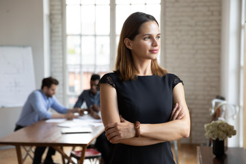 Businesswoman standing with arms crossed looks in window daydreaming about career growth, plan...