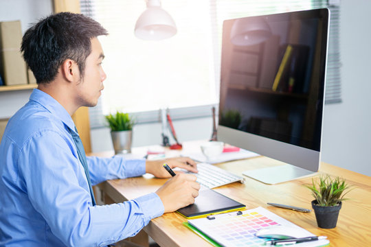 asian office worker using graphics tablet with a pen digital drawing or designing looking at the monitor screen, working in an office environment with clipboard of colour charts on computer desk