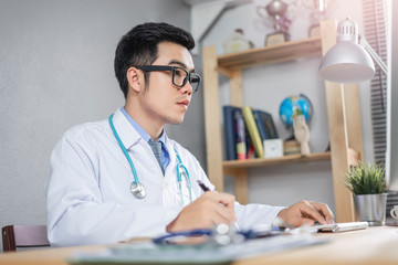 asian male medical doctor smiling using a pen writing signing a health insurance form or contract, wearing a lab coat sitting in his hospital office with stethoscope and computer on the wooden desk