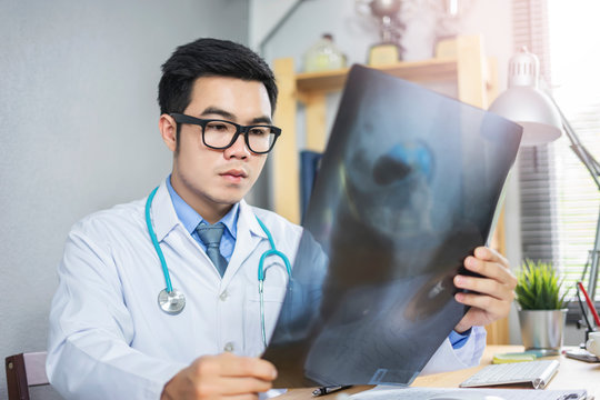 male asian medical doctor looking and examining a bone structure of an x-ray filter, holding the filter and diagnosing his patient health, wearing a lab coat stethoscope working in a hospital office