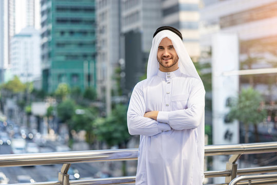 A Caucasian Arab man standing on a bridge platform with arms across the chest posture, wearing rational arab garment disdasha and thobe, within the city center, nature and busy road in the background