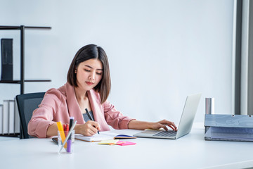 Obraz premium beautiful female asian worker using laptop computer typing on keyboard and writing on notebook, work in an office with folder files sticky notes for ideas information, wearing pink formal clothing.