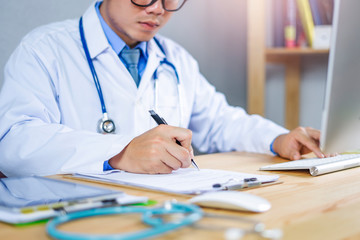 asian male medical doctor smiling using a pen writing signing a health insurance form or contract, wearing a lab coat sitting in his hospital office with stethoscope and computer on the wooden desk