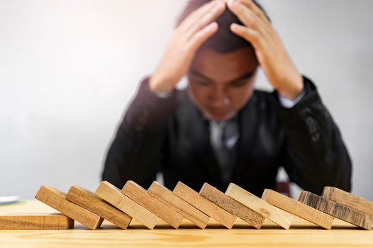 Concept Of A Male Asian Businessman Covering His Head Looking Down With Hands Falling Stress Sad, With Fallen Wooden Dominos Representing Fail And Business Going Bankrupt Within The Business Industry