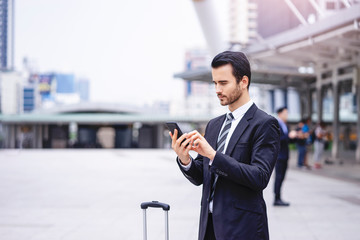Caucasian businessman standing confidently  smart phone device by holding and touching the screen, wearing formally black suit and tie with tall city skylines and metro station in the background..