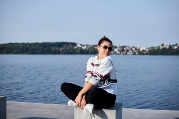 Sep 7, 2019-Ternopil/Ukraine:Young brunette woman, wearing black leggings and white sweatshot, sitting, resting on concrete platform by city lake with bicycle aside. Sports bike ride in summer morning