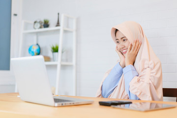 Pretty young Asian woman Muslim with turban hijab using laptop at a wooden table in the day to search and doing office work in an office, business, finance technology work station concept