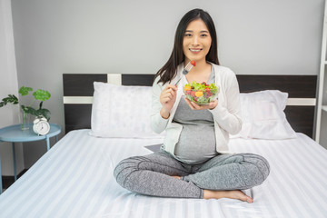 Obraz na płótnie Canvas Beautiful asian pregnant woman holding a bowl of salad of vegetables and fruits, using a fork eating a strawberry. Eating healthy having lunch sitting on the white bed, relaxing and resting in bedroom