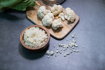 Cauliflower rice in a wooden bowl with some cauliflower pieces in background. Close up view.