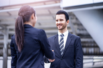 caucasian businessman smiling and standing casually shaking hands with a beautiful asian businesswoman in greetings, outdoor within the city district with urban architectural structure in background