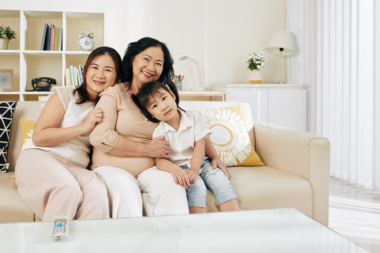 Happy Vietnamese serious woman with her adult daughter and little granddaughter sitting on sofa at home