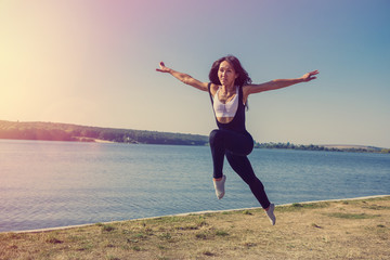 Fototapeta na wymiar Smiling young female gymnast is jumping in split outdoors near the lake. Healthy lifestyle