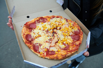 Fresh appetizing pizza with corn salami and double cheese in girl's hands. Pizza in the box outdoor. Meat pizza with ham.