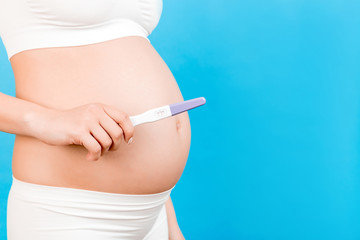 Close up of positive pregnancy test against pregnant woman's belly at blue background. Future mother in white underwear. Baby expecting. Copy space