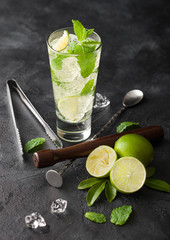 Glass of Mojito cocktail with ice cubes mint and lime on black board with spoon and muddler and fresh limes. Sparkling refreshing water.