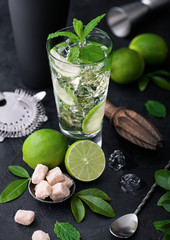 Glass of Mojito cocktail with ice cubes mint and lime on black board with spoon and fresh limes and cane sugar with wooden squeezer and black shaker with ice.