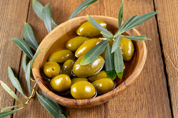 Close up of green olives in olive oil and an olive branch in a small olive wood bowl on a rustic...
