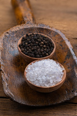 Black whole peppercorns and blue crystal salt in wooden bowls on an old flour shovel on a rustic wooden background