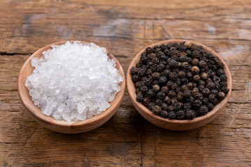 Coarse crystal salt and whole black peppercorns in small wooden bowls on a rustic wooden background