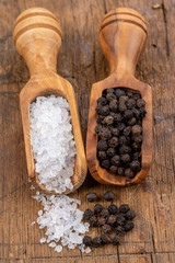 Close-up of coarse crystal salt and whole black peppercorns on small spice shovels made of olive wood on a rustic wooden background