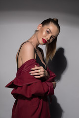Fototapeta premium A beautiful young woman with red lips and a high tail in a Burgundy raincoat and a black bra. Fashion art Studio portrait