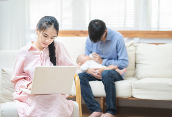Fototapeta premium Young asian mother using laptop computer in living room at home with blurred of father acting mom feeding milk bottle in background his son baby 1 year old, work at home and social distancing concept.