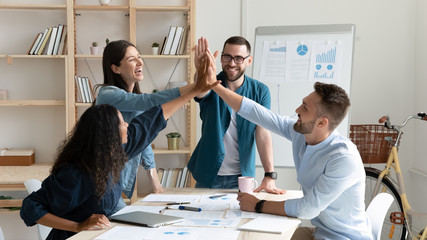 Overjoyed diverse businesspeople gather in office gave high five celebrate shared victory or win,...