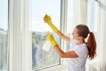 Woman in white shirt and yellow rubber gloves cleaning window with cleanser sprayer and yellow rag at home or office, copy space. People, housework and housekeeping concept