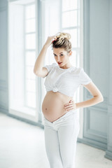 A pregnant European girl stands by the window. Studio photo shoot. Soft focus.