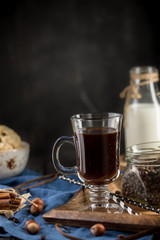 Cup of coffee with coffee beans, spoon, cinnamon sticks, bottle of milk and cookies on black background