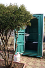 Fototapeta na wymiar Cabin of a plastic biotoilet with an open door behind a juniper bush in the country