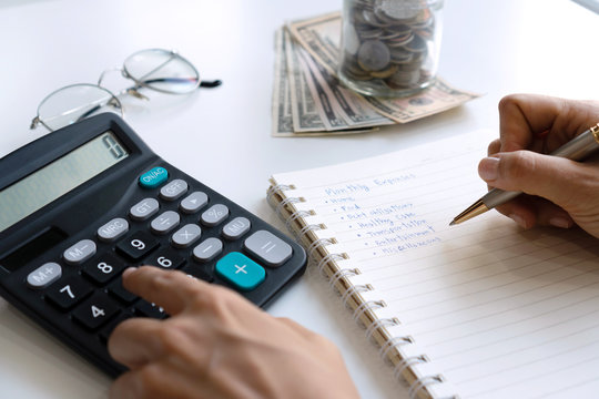 Woman writing home monthy expenses in notebook while using calculator on desk. Copy space, close up.