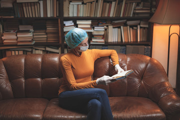 Obraz premium Woman with protective antiviral mask sitting at home in isolation / quarantine and reading book.