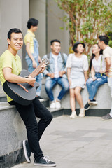 Creative young Vietnamese student standing on campus of colleage of art and playing guitar
