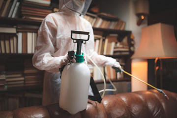 Person with protective antiviral mask, chemical decontamination sprayer bottle disinfecting household and furniture.