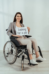 Obraz na płótnie Canvas Disabled woman smiling and holding placard with looking for a job lettering on wheelchair on white