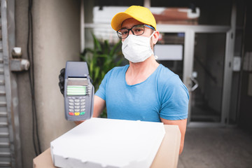 Deliveryman with protective medical mask holding pizza box and POS wireless terminal for card paying - days of viruses and pandemic, food delivery to your home and safety hygiene measures.