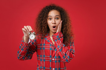 Shocked young african american girl in pajamas homewear posing resting at home isolated on red background. Relax good mood lifestyle concept. Mock up copy space. Hold alarm clock, put hand on cheek.