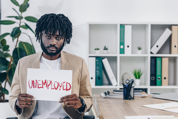 African american employee showing placard with unemployed lettering in office - obrazy, fototapety, plakaty
