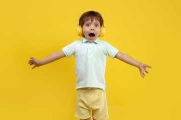 Shocked little boy in casual outfit listening music in headphone and stretching out arms over yellow background.