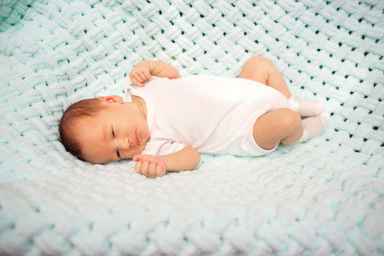 A newborn baby in a white bodysuit lies in his cradle on a blue knitted blanket