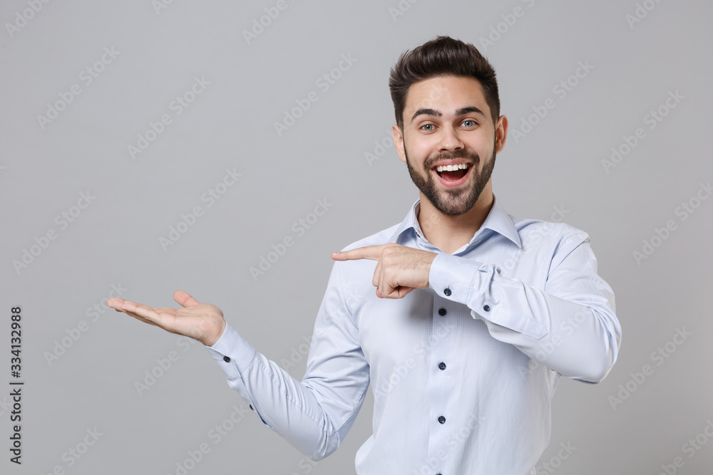 Canvas Prints Cheerful young unshaven business man in light shirt posing isolated on grey wall background in studio. Achievement career wealth business concept. Mock up copy space. Pointing index finger hand aside.