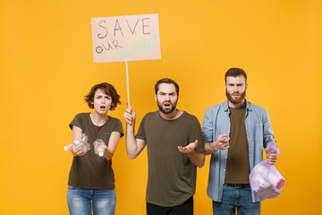 Puzzled protesting young three people hold protest broadsheet placard plastic bottles, trash bag isolated on yellow background. Stop nature garbage ecology environment protection concept. Save planet.