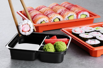 Sushi maki with shrimp and avocado, sushi philadelphia, soy sauce, wasabi and ginger in a take away plastic container. Chopsticks taking portion of sushi roll.