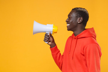 Side view of smiling young african american man guy in red streetwear hoodie posing isolated on yellow background studio portrait. People emotion lifestyle concept. Mock up copy space. Hold megaphone.