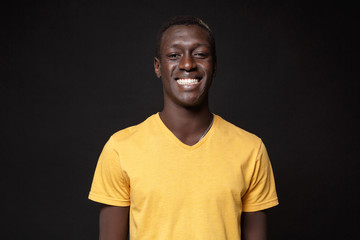 Smiling young african american man guy in yellow t-shirt posing isolated on black wall background studio portrait. People sincere emotions, lifestyle concept. Mock up copy space. Looking camera.