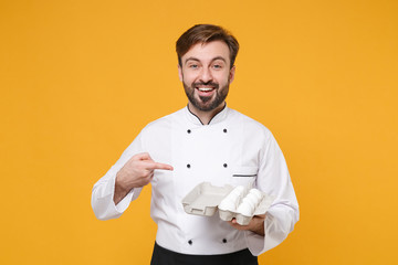 Cheerful young bearded male chef cook or baker man in white uniform shirt posing isolated on yellow background in studio. Cooking food concept. Mock up copy space. Pointing index finger on egg tray.