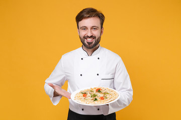 Smiling young bearded male chef cook or baker man in white uniform shirt isolated on yellow background in studio. Cooking food concept. Mock up copy space. Pointing hand on italian pizza on plate.