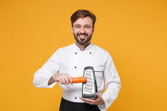 Smiling young bearded male chef cook or baker man in white uniform shirt posing isolated on yellow background studio portrait. Cooking food concept. Mock up copy space. Holding grater and carrots.