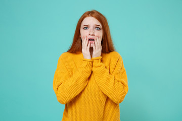 Shocked worried young redhead woman girl in yellow sweater posing isolated on blue turquoise background studio portrait. People emotions lifestyle concept. Mock up copy space. Put hands on cheeks.